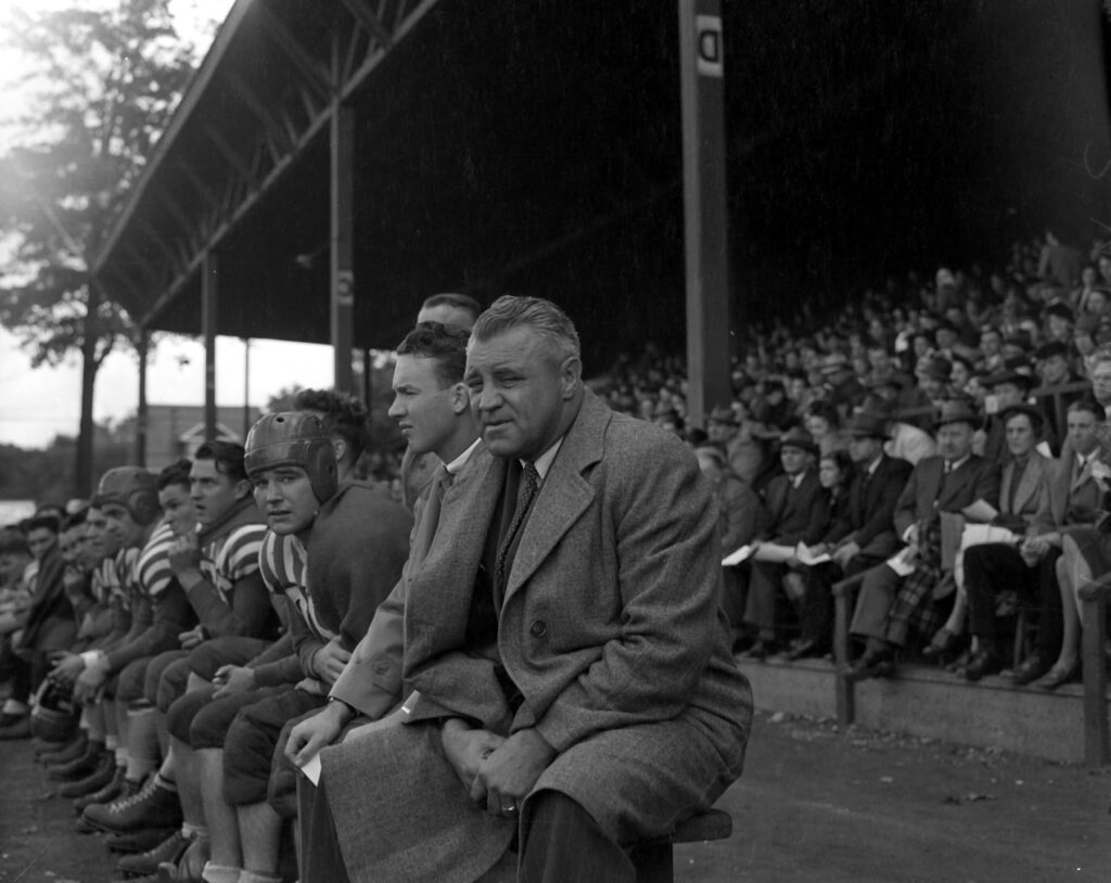 Brian Timmis sits on a football bench with his team. A large crowd is seated behind him.