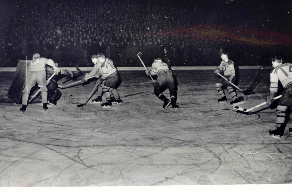 Seven ice hockey players surround the goalie on a hockey rink. They're playing for gold in the 1948 Olympic Winter Games in St. Moritz, Switzerland.