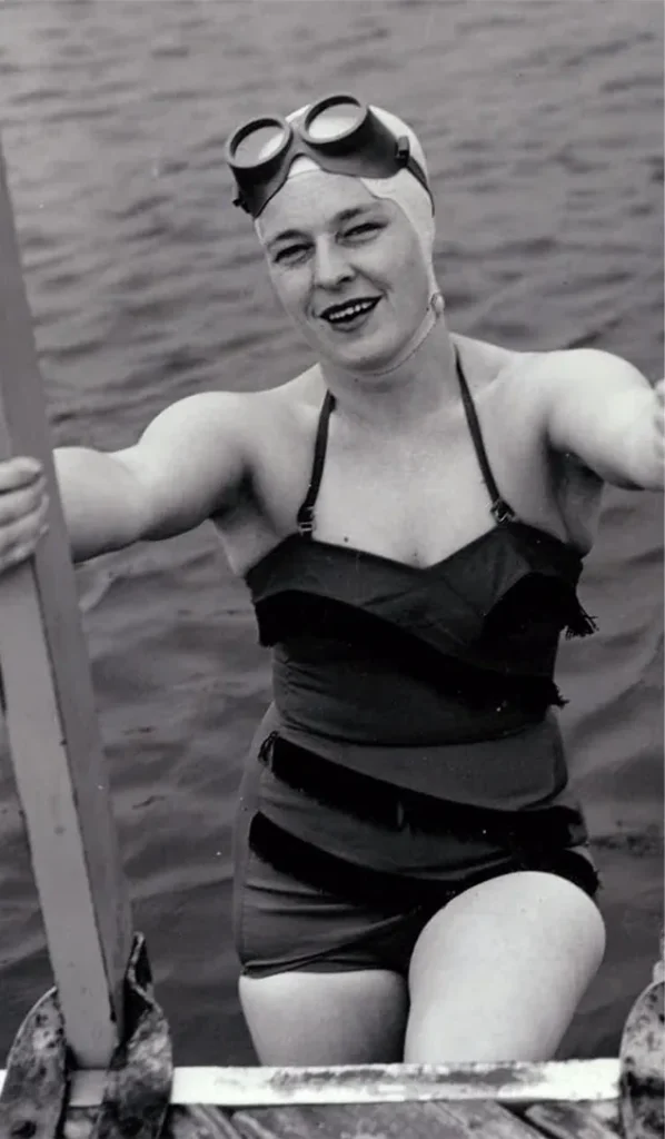 Black and white, Winnie Roach getting out of the water. She's wearing a one-piece bathing suit, swim googles, and a swim cap.